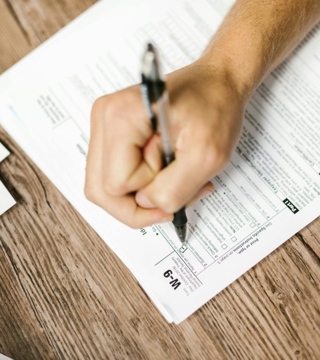 Hand writing on a W-9 tax form using a ballpoint pen on wooden desk.